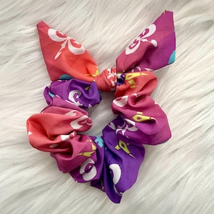 May include: A colorful scrunchie with a bow, featuring a floral pattern in shades of pink, purple, and orange. The scrunchie is made of fabric and is designed to hold hair. The bow adds a decorative element to the hair accessory.