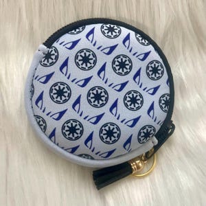 Op de afbeelding: Een ronde, witte muntbeurs met een blauw en zwart patroon van sterren en vleugels. De beurs heeft een zwarte kwast en een gouden rits.