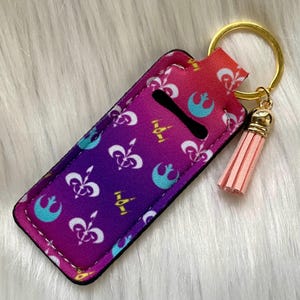 May include: A fabric lip balm holder with a gold keychain and a pink tassel. The holder features a purple to red gradient with white fleur-de-lis, teal rebel alliance symbols, and gold spaceship graphics.