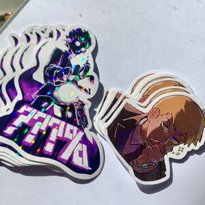 Puede incluir: Dos pegatinas holográficas con personajes de la serie de anime Mob Psycho 100. La primera pegatina es morada y presenta un personaje con cabello negro y un orbe blanco en su mano. El texto "MOB PSYCHO 100" está escrito en letras blancas en la pegatina. La segunda pegatina es marrón y presenta un personaje con cabello rubio y una expresión seria. El personaje lleva un traje oscuro y una corbata.