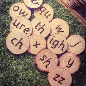 May include: A collection of circular wooden slices featuring dark, burned-in letters and letter combinations including ear, qu, ow, ure, th, w, ch, x, igh, j, sh, y, and zz. Each piece displays natural wood grain patterns and bark edges.