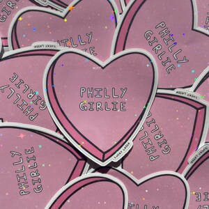 May include: A collection of pink heart-shaped stickers with the text "PHILLY GIRLIE" in a bold font. Each sticker has a white border and a pink outline. The stickers are scattered across a surface, with a holographic glitter effect.