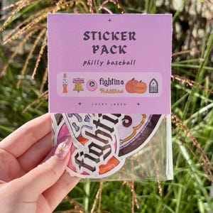 May include: A sticker pack in a clear plastic sleeve with a light purple header card. The card features the text STICKER PACK, philly baseball, and LUCKY LASSO. The stickers include graphics of a lava lamp, a bell, pumpkins, and the text fightins.
