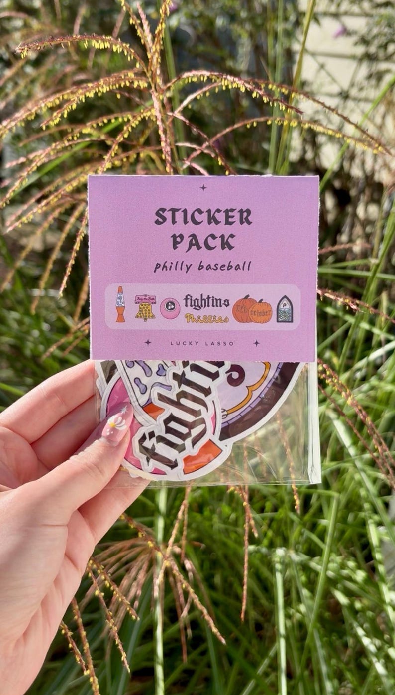 May include: A sticker pack in a clear plastic sleeve with a light purple header card. The card features the text STICKER PACK, philly baseball, and LUCKY LASSO. The stickers include graphics of a lava lamp, a bell, pumpkins, and the text fightins.