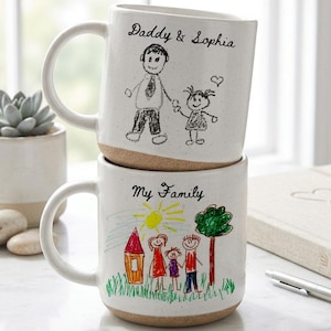 May include: Two stacked white ceramic mugs with drawings and text. The top mug says "Daddy & Sophia" with a drawing of a father and child. The bottom mug says "My Family" with a drawing of a family. Both mugs have a tan base.