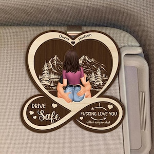 Può includere: Accessorio per auto a forma di cuore in legno con illustrazione di coppia e paesaggio montano. L'accessorio riporta i nomi "Olivia" e "Hudson" e le frasi "Drive Safe" e "Fucking Love You (either way works)".