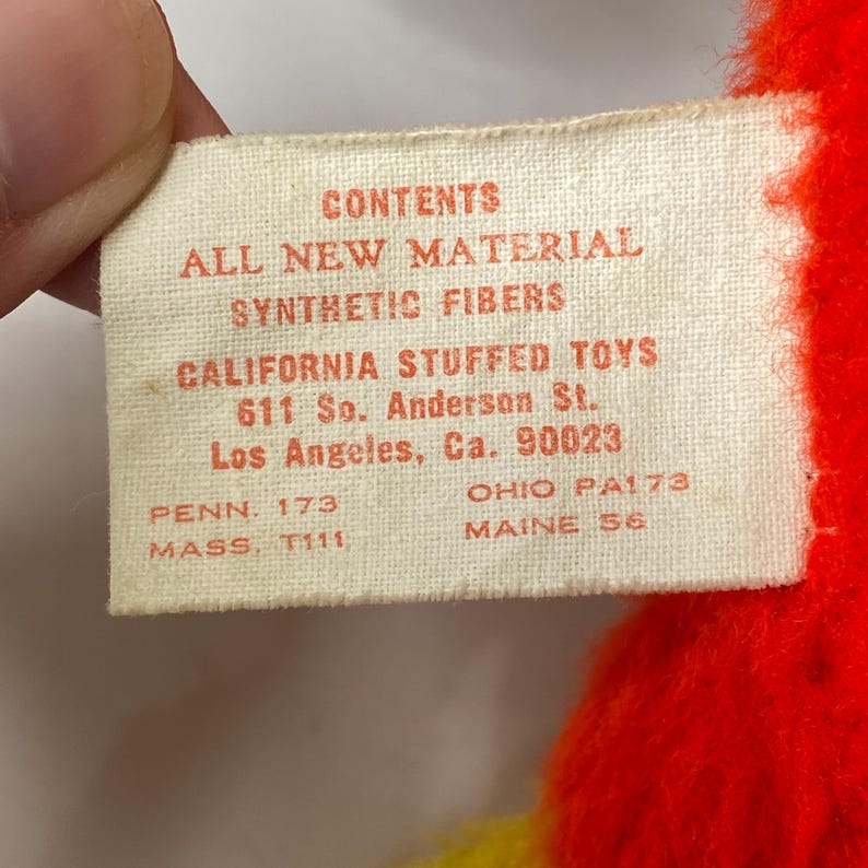 Puede incluir: Primer plano de una etiqueta crema con texto rojo. La etiqueta detalla el contenido de un juguete de peluche, incluyendo "ALL NEW MATERIAL" y "SYNTHETIC FIBERS". Tambi&eacute;n enumera el fabricante, "CALIFORNIA STUFFED TOYS", y una direcci&oacute;n en Los &Aacute;ngeles. Tambi&eacute;n son visibles c&oacute;digos de estado adicionales.