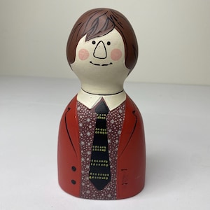 May include: A ceramic figurine of a person with brown hair, a white face, and rosy cheeks. The figure wears a red jacket over a shirt with a star pattern and a black tie with yellow and white dots. The figurine is a decorative item.