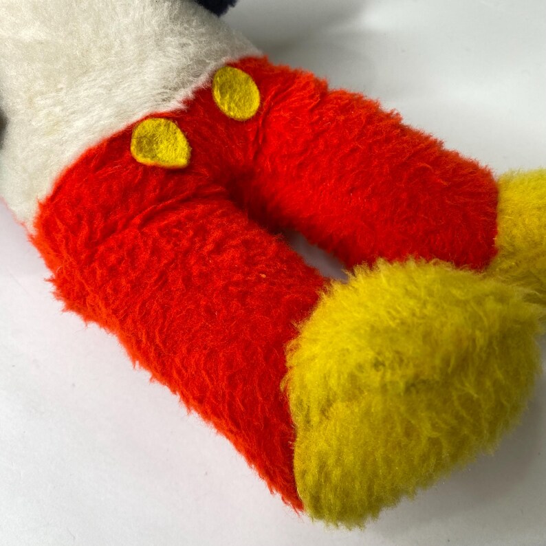 Puede incluir: Primer plano de las piernas y los pies de un peluche. Las piernas son de color rojo brillante con dos botones amarillos, y los pies son amarillos. El torso blanco y los pantalones cortos negros del juguete son visibles en el fondo.