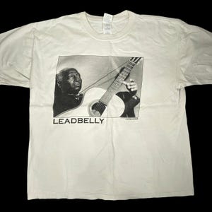 May include: Off-white t-shirt featuring a black and white image of Leadbelly playing a guitar. The image includes the text "LEADBELLY" in a bold font at the bottom of the image. The shirt has a crew neck and short sleeves.