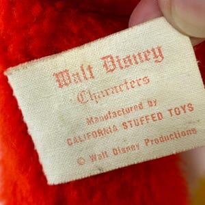 Puede incluir: Primer plano de una etiqueta vintage de un peluche de personaje de Walt Disney. La etiqueta es rectangular, con texto rojo sobre fondo crema. Dice "Walt Disney Characters" y "Manufactured by CALIFORNIA STUFFED TOYS".