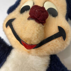 Puede incluir: Primer plano de un juguete de peluche vintage con una cara sonriente. El juguete presenta una cara de color crema, orejas azul oscuro y una nariz burdeos. Los ojos son negros con reflejos blancos y la boca est&aacute; delineada en negro con una sonrisa roja.