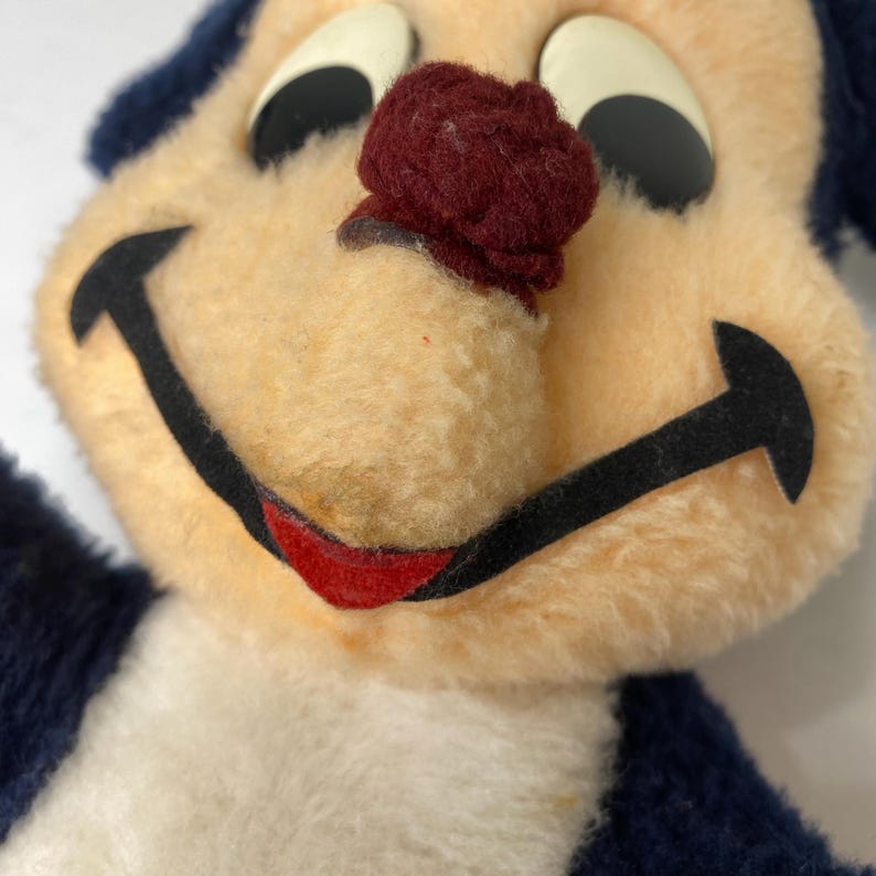 Puede incluir: Primer plano de un juguete de peluche vintage con una cara sonriente. El juguete presenta una cara de color crema, orejas azul oscuro y una nariz burdeos. Los ojos son negros con reflejos blancos y la boca est&aacute; delineada en negro con una sonrisa roja.