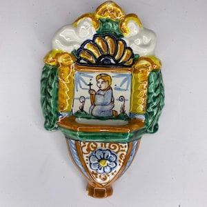 Può includere: Un'acquasantiera in ceramica colorata da parete. Presenta una figura inginocchiata con una croce, con una parte superiore a forma di conchiglia e dettagli floreali e fogliari. La tavolozza dei colori include blu, verde, giallo e marrone.
