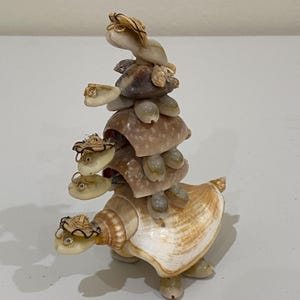 May include: A decorative figurine crafted from various seashells, featuring a vertical stack of four turtle-like creatures. Each creature has a small woven hat, wire-rimmed glasses, and a shell body. The base is a large, light-colored conch shell.