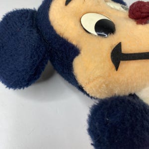 Puede incluir: Primer plano de un juguete de peluche vintage con la cara de un personaje de dibujos animados. El juguete tiene la cabeza y las orejas de color azul marino, la cara de color canela y un ojo en blanco y negro. La boca es una forma negra curvada y la nariz es un peque&ntilde;o pomp&oacute;n rojo.