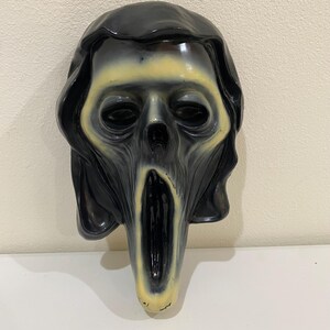 May include: A molded plastic ghost mask featuring a glossy black hood and a pale, yellowish-white face. The mask has hollow, dark eye sockets and an elongated, open mouth, creating a distorted, screaming expression with smooth, reflective surfaces.
