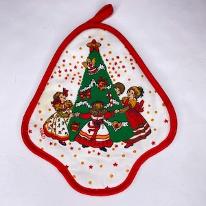 Puede incluir: Manopla blanca con temática navideña, con ribete y lazo para colgar rojos. El diseño presenta un árbol de Navidad verde decorado con lazos y ángeles, rodeado de niños con vestidos festivos. Estrellas y puntos dorados completan la escena.