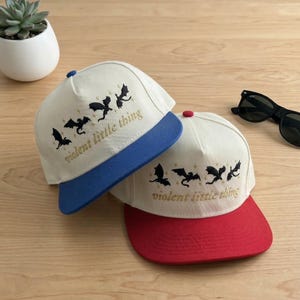 May include: Two five-panel baseball caps with off-white crowns and flat brims. One cap has a blue brim, the other a red brim. Both feature black embroidered dragon silhouettes, gold star accents, and the text "violent little thing" in gold.
