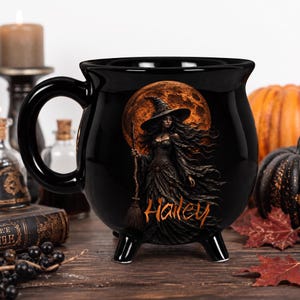 May include: A black ceramic cauldron-shaped mug featuring a graphic of a witch in a pointed hat holding a broomstick against a large orange full moon. The name Hailey is printed in orange script lettering at the bottom of the graphic design.