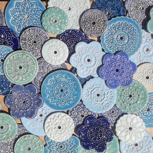 Könnte beinhalten: Eine Auswahl an dekorativen Keramikteilen in verschiedenen Blau-, Weiß- und Türkistönen. Die Teile sind rund, blütenförmig und andere geometrische Designs, jedes mit einem zentralen Loch und geprägten Mustern. Die Teile sind eng aneinander angeordnet.