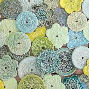 Könnte beinhalten: Eine Sammlung handgefertigter Keramikuntersetzer in verschiedenen Formen und Pastellfarben, darunter Blau, Grün und Gelb. Jeder Untersetzer hat ein einzigartiges Blumen- oder Mandala-Design mit einem zentralen Loch.