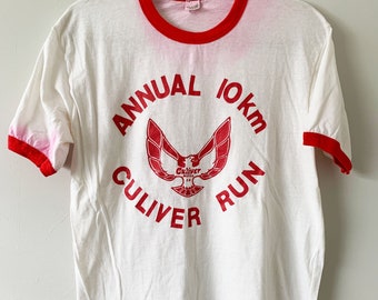 May include: White t-shirt with a red ringer and a red graphic of a winged bird with the text "Annual 10km Culver Run".