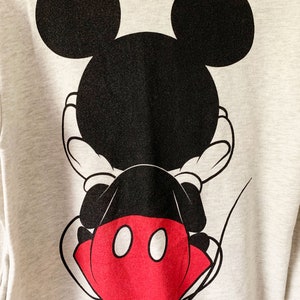 Puede incluir: Una sudadera gris con un gr&aacute;fico de Mickey Mouse negro y rojo en la espalda. Mickey Mouse est&aacute; de espaldas al espectador con las manos detr&aacute;s de la espalda.