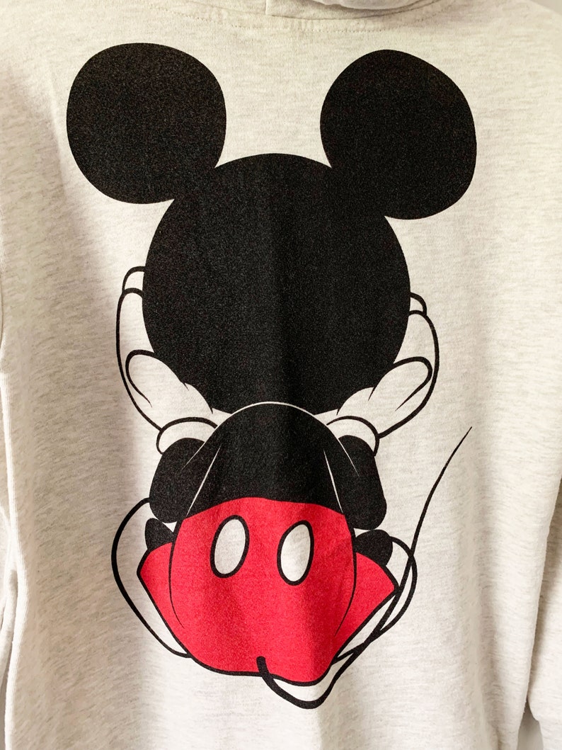Puede incluir: Una sudadera gris con un gr&aacute;fico de Mickey Mouse negro y rojo en la espalda. Mickey Mouse est&aacute; de espaldas al espectador con las manos detr&aacute;s de la espalda.