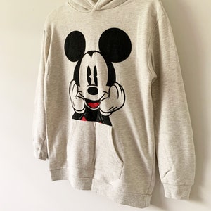 Puede incluir: Una sudadera con capucha gris con un gr&aacute;fico de Mickey Mouse en blanco y negro en la parte delantera. La sudadera tiene un bolsillo canguro.