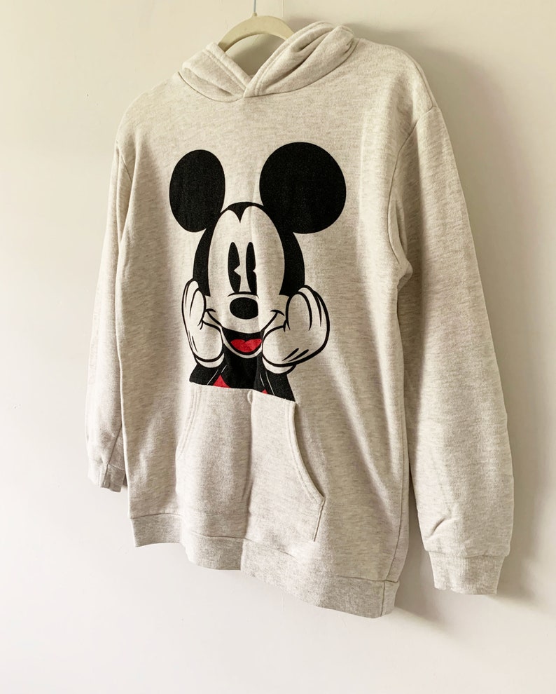 Puede incluir: Una sudadera con capucha gris con un gr&aacute;fico de Mickey Mouse en blanco y negro en la parte delantera. La sudadera tiene un bolsillo canguro.