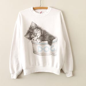 Op de afbeelding: Witte sweatshirt met een gedetailleerde illustratie van een slapende grijze kat. De kat zit in een bak met het woord "DOG" in lichtblauw. De sweatshirt heeft lange mouwen en een ronde hals.