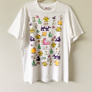 May include: White t-shirt with a colorful cartoon print of various animals, including dogs, cats, pigs, and a dragon. The animals are all in different poses and expressions, and the print covers the entire front of the shirt.