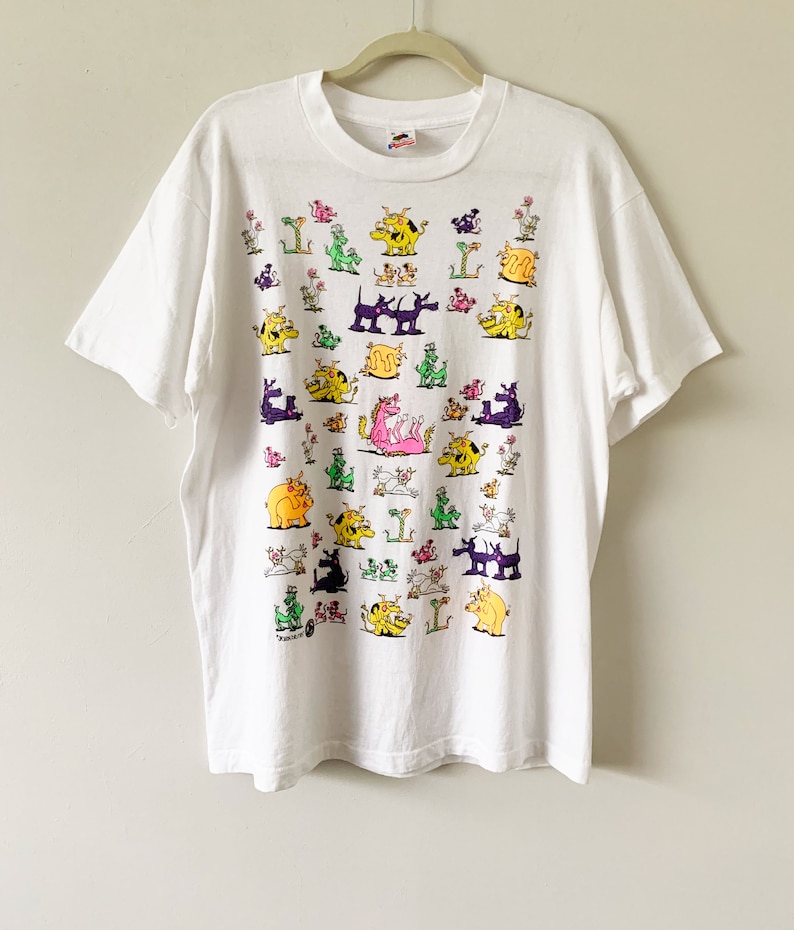 May include: White t-shirt with a colorful cartoon print of various animals, including dogs, cats, pigs, and a dragon. The animals are all in different poses and expressions, and the print covers the entire front of the shirt.