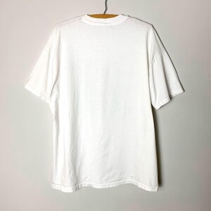 May include: A plain white t-shirt hanging on a wooden hanger. The short-sleeved shirt is made of a soft, opaque fabric. The shirt is viewed from the back, showing the neckline and the shoulders.