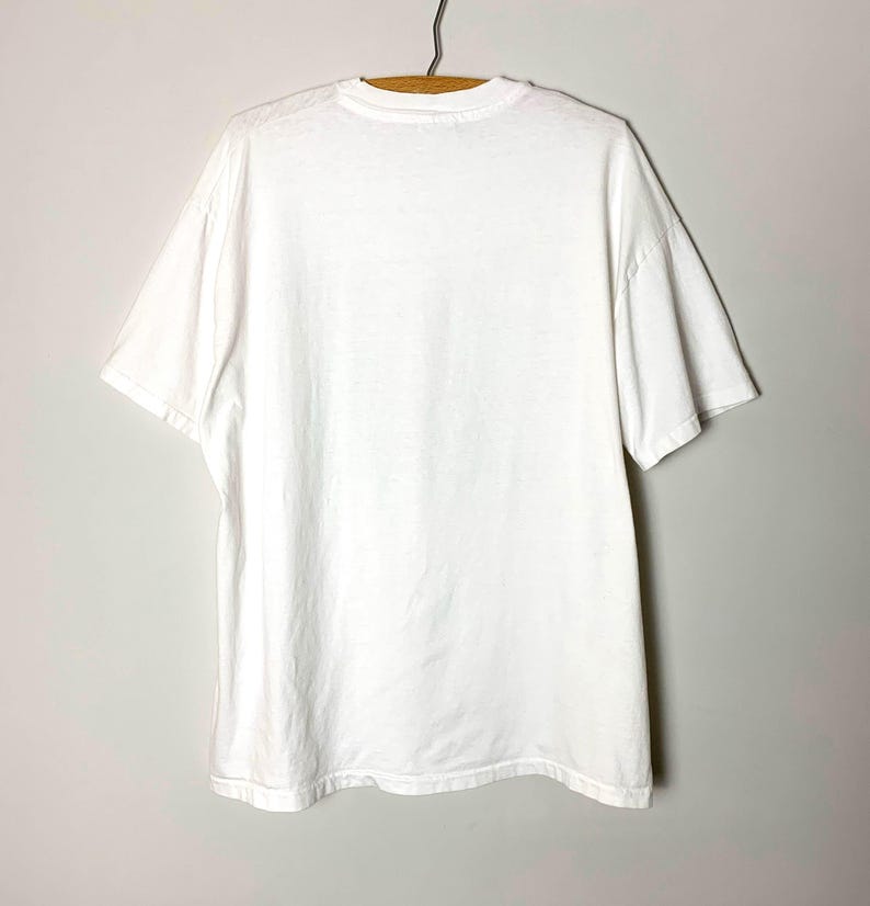 May include: A plain white t-shirt hanging on a wooden hanger. The short-sleeved shirt is made of a soft, opaque fabric. The shirt is viewed from the back, showing the neckline and the shoulders.