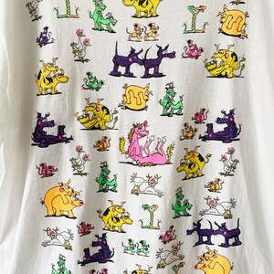 May include: White t-shirt with a colorful graphic print of various animals in different positions. The animals include pigs, cows, dogs, snakes, and birds. The print is a repeating pattern and covers the entire shirt.
