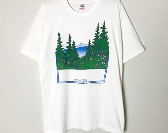 May include: White t-shirt with a nature scene featuring green trees, a mountain, and wildflowers. The word "OREGON" is printed in blue at the bottom of the graphic. The shirt is hanging on a wooden hanger.