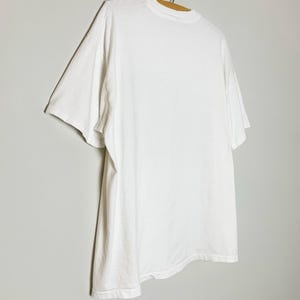 May include: A plain white t-shirt hanging on a wooden hanger. The short-sleeved shirt is made of a soft, opaque fabric. The shirt is hanging against a light gray background.