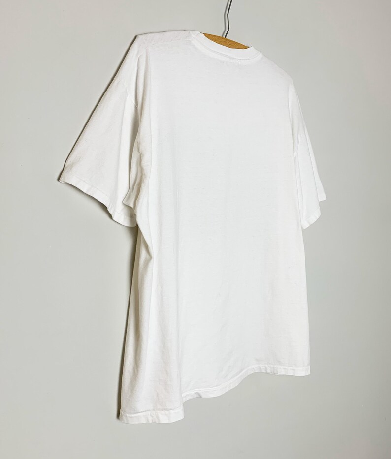 May include: A plain white t-shirt hanging on a wooden hanger. The short-sleeved shirt is made of a soft, opaque fabric. The shirt is hanging against a light gray background.