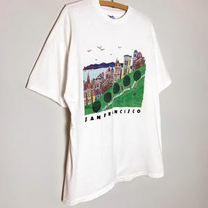 May include: White t-shirt featuring a colorful illustration of San Francisco's cityscape. The design includes buildings, trees, and the text "SAN FRANCISCO" in black. The shirt is hanging on a wooden hanger.