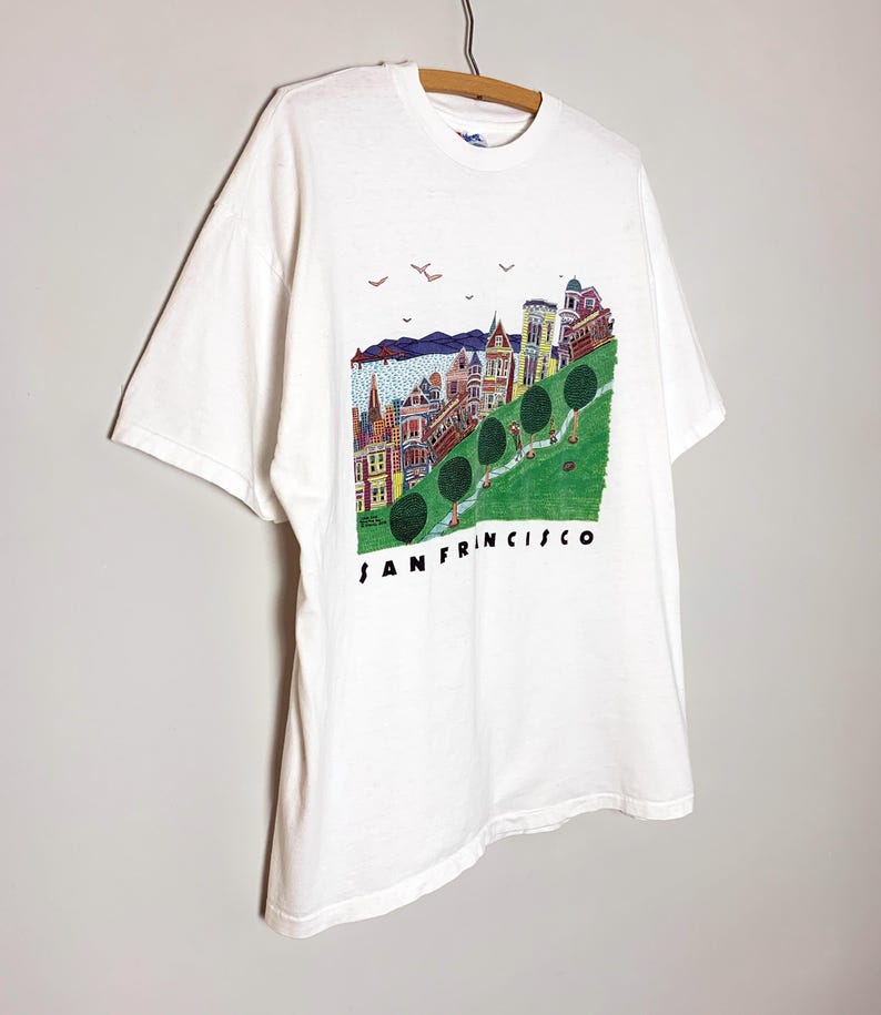 May include: White t-shirt featuring a colorful illustration of San Francisco's cityscape. The design includes buildings, trees, and the text "SAN FRANCISCO" in black. The shirt is hanging on a wooden hanger.