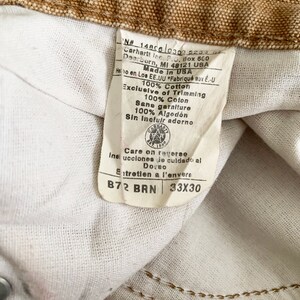 Puede incluir: Una etiqueta blanca con texto marr&oacute;n y un logotipo marr&oacute;n de Carhartt. La etiqueta dice "N# 1480603005230 Carhartt Inc. Box 600 Dearborn, MI 48121 USA Made in USA 100% Cotton Exclusive of Trimming 100% Coton Sans garniture 100% Algod&oacute;n Sin incluir adorno Care on reverse Insi ucciones de culdado al Dovno Entretien a l'envers B72 BRN 33X30"