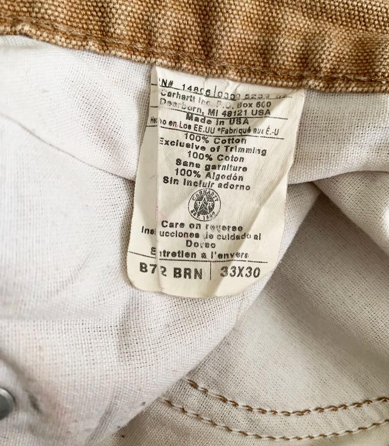 Puede incluir: Una etiqueta blanca con texto marr&oacute;n y un logotipo marr&oacute;n de Carhartt. La etiqueta dice "N# 1480603005230 Carhartt Inc. Box 600 Dearborn, MI 48121 USA Made in USA 100% Cotton Exclusive of Trimming 100% Coton Sans garniture 100% Algod&oacute;n Sin incluir adorno Care on reverse Insi ucciones de culdado al Dovno Entretien a l'envers B72 BRN 33X30"
