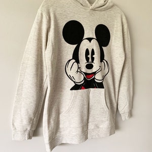 Puede incluir: Una sudadera con capucha de color gris claro con un gr&aacute;fico de Mickey Mouse en blanco y negro en la parte delantera. Mickey Mouse est&aacute; sonriendo y tiene las manos levantadas hacia su cara. La sudadera tiene un bolsillo canguro.