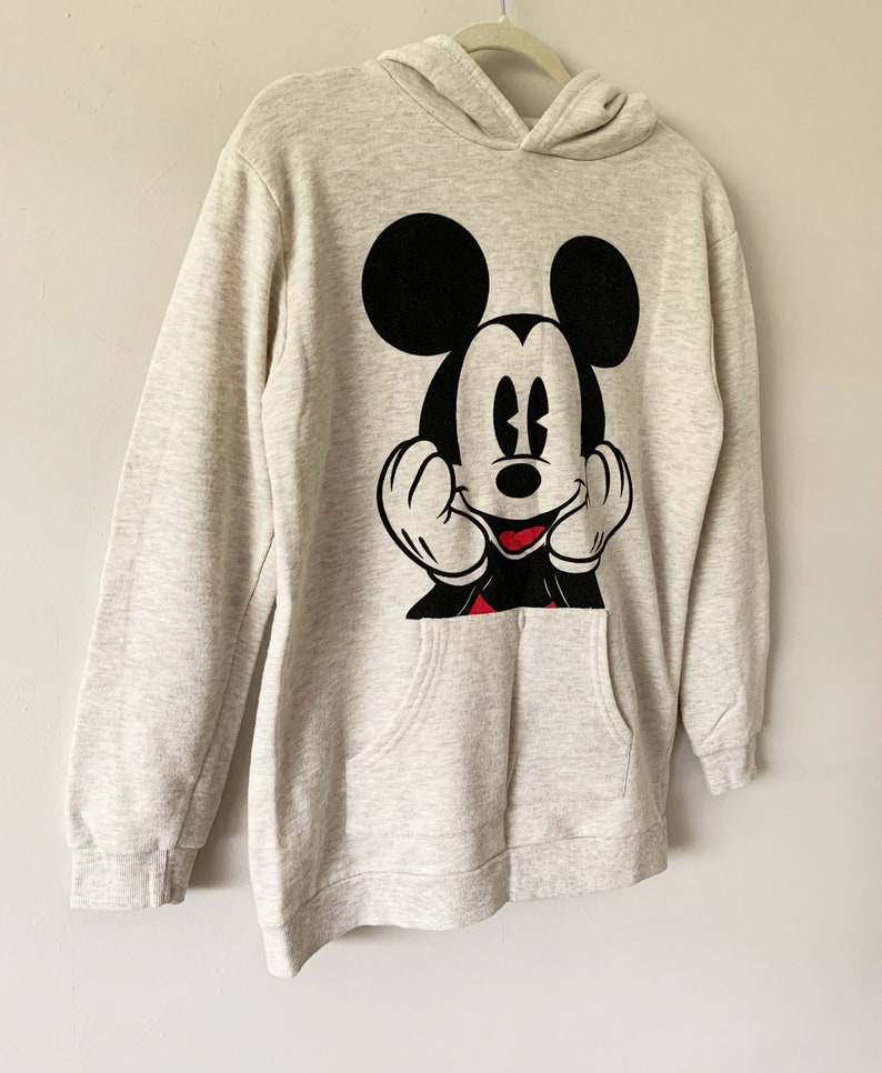 Puede incluir: Una sudadera con capucha de color gris claro con un gr&aacute;fico de Mickey Mouse en blanco y negro en la parte delantera. Mickey Mouse est&aacute; sonriendo y tiene las manos levantadas hacia su cara. La sudadera tiene un bolsillo canguro.