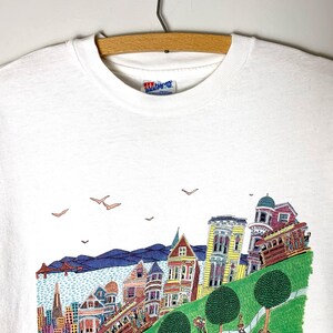 May include: White t-shirt featuring a colorful illustration of San Francisco's iconic landmarks, including the Golden Gate Bridge, cable cars, and Victorian houses. The design includes blue, yellow, red, and green elements, with birds in the sky.