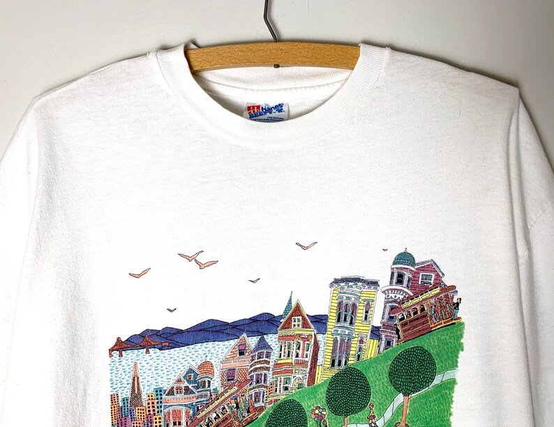 May include: White t-shirt featuring a colorful illustration of San Francisco's iconic landmarks, including the Golden Gate Bridge, cable cars, and Victorian houses. The design includes blue, yellow, red, and green elements, with birds in the sky.