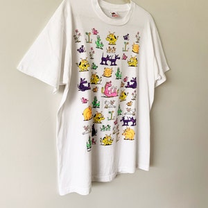May include: White t-shirt with a colorful cartoon print of various animals, including dogs, cats, and a dragon. The print is all over the shirt and features a variety of colors, including pink, yellow, green, and purple.