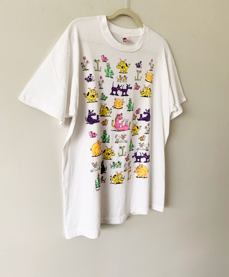 May include: White t-shirt with a colorful cartoon print of various animals, including dogs, cats, and a dragon. The print is all over the shirt and features a variety of colors, including pink, yellow, green, and purple.
